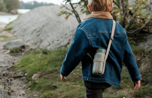 Child in a denim jacket with a backpack walking outdoors near a body of water.