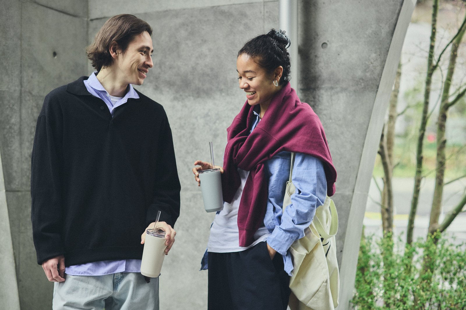 Two people standing outdoors, holding drinks, with a concrete wall and greenery in the background.